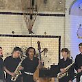 Fünf jugendliche Musiker stehen auf einer Bühne mit gefliester Wand und präsentieren ihr Ensemble, bestehend aus zwei Saxofonisten, einem Kontrabassisten und zwei weiteren Mitgliedern. Die Gruppe, gekleidet in überwiegend dunkle Kleidung, tritt in einem historisch wirkenden Veranstaltungsraum auf.
