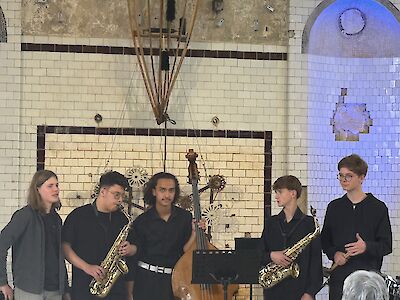 Fünf jugendliche Musiker stehen auf einer Bühne mit gefliester Wand und präsentieren ihr Ensemble, bestehend aus zwei Saxofonisten, einem Kontrabassisten und zwei weiteren Mitgliedern. Die Gruppe, gekleidet in überwiegend dunkle Kleidung, tritt in einem historisch wirkenden Veranstaltungsraum auf.