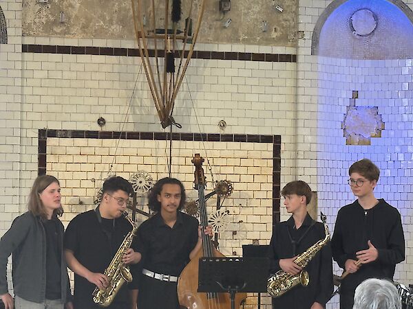 Fünf jugendliche Musiker stehen auf einer Bühne mit gefliester Wand und präsentieren ihr Ensemble, bestehend aus zwei Saxofonisten, einem Kontrabassisten und zwei weiteren Mitgliedern. Die Gruppe, gekleidet in überwiegend dunkle Kleidung, tritt in einem historisch wirkenden Veranstaltungsraum auf.